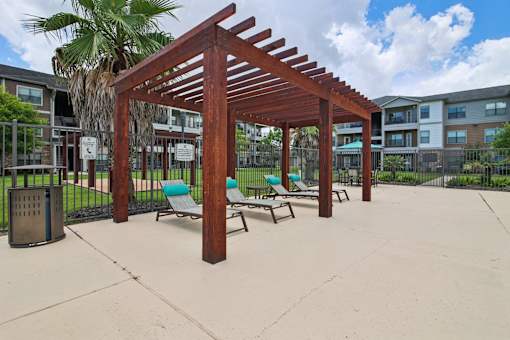 A wooden pergola with blue chairs and a trash can in front of apartment buildings.