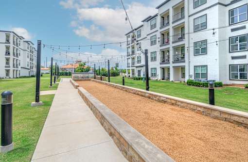 A long concrete walkway leads to apartment buildings with a grassy area and string lights on the side.