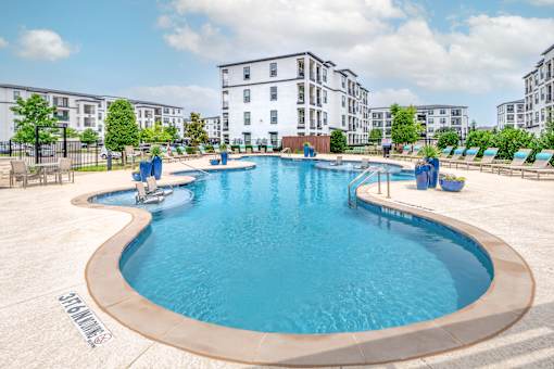 A large outdoor swimming pool surrounded by lounge chairs and buildings in the background.