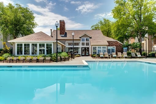 A large swimming pool in front of a house with a patio and chairs.