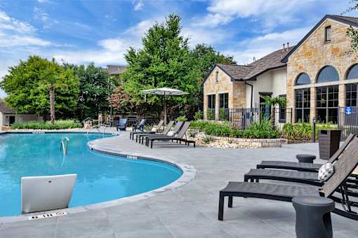 Pool deck with chaise lounges beside a stone-style clubhouse exterior and gardens.