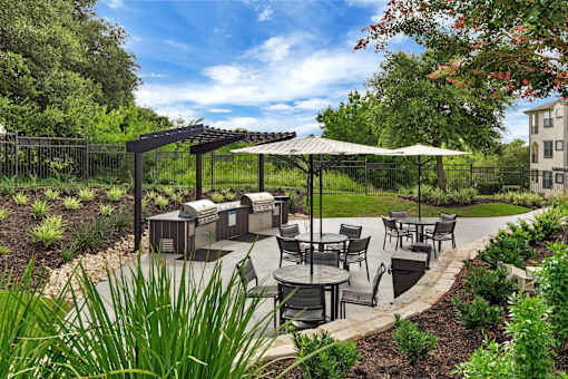 Landscaped patio with grills, outdoor dining tables, and shaded seating near greenery.