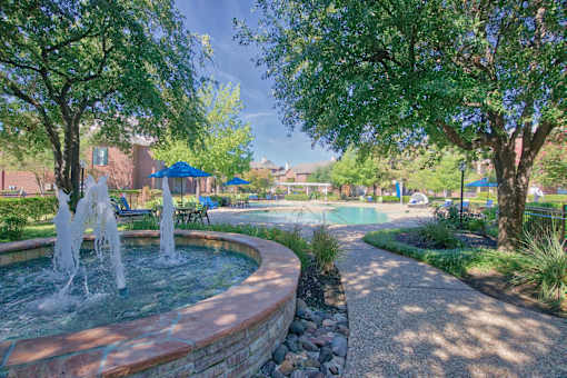 A fountain in the middle of a park with trees and a pool in the background.