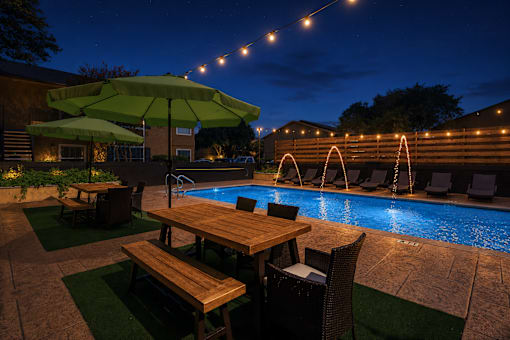 A poolside table with chairs and umbrellas is lit up at night.