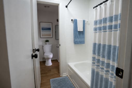 A white toilet is in a small bathroom with a blue rug and a white tub.