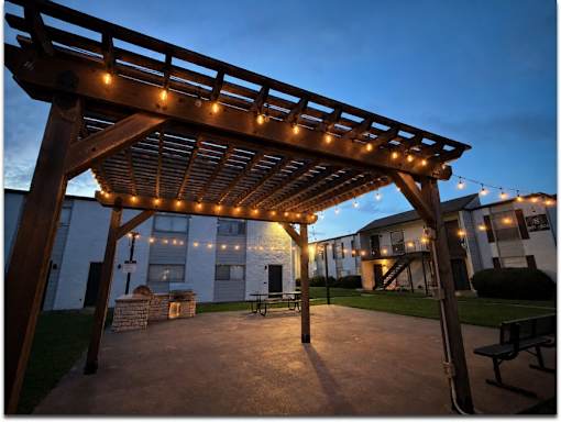 A wooden pergola with string lights is in the foreground of a residential area.