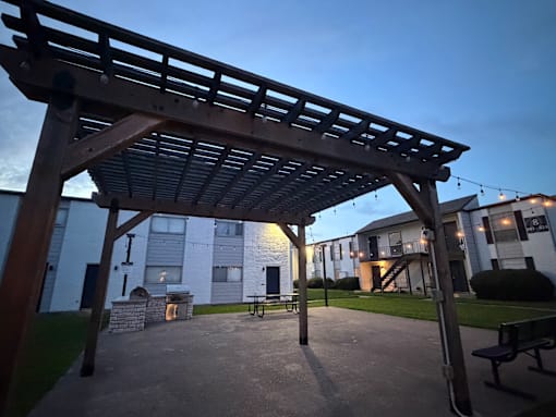 A wooden pergola structure is in the foreground of a park-like setting with benches and a building in the background.