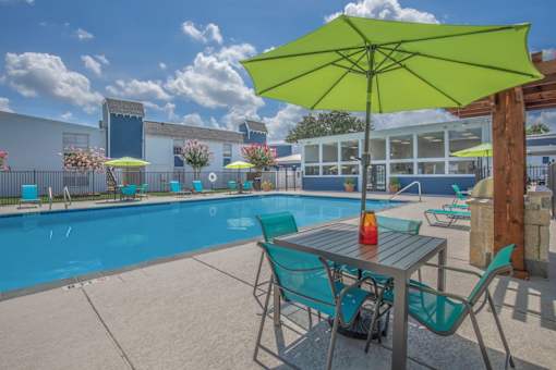 A table with a yellow umbrella is in the foreground of a pool area.