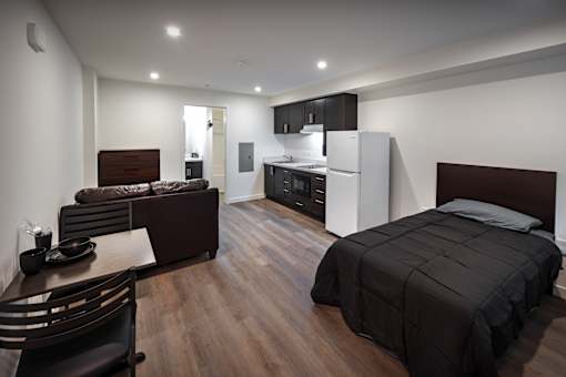 A modern bedroom with a large bed and a kitchen area in the background.