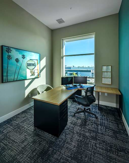 A room with a desk and a chair with a painting of a surfing scene on the wall.