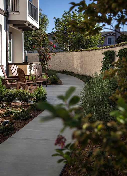 A concrete walkway leads to a house with a patio and chairs.