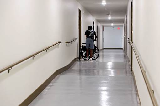 A man in a black shirt and blue shorts is pushing a wheelchair down a long hallway with white walls and a reflective floor.