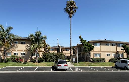 A parking lot with a white van and a silver car parked in front of apartment buildings.