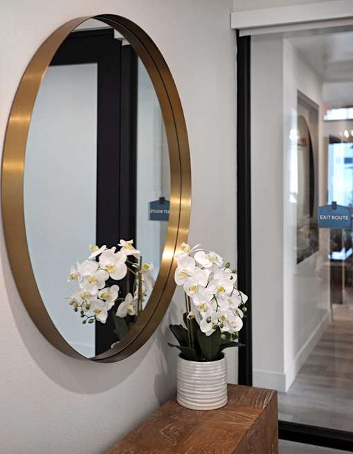A round mirror with a gold frame is on a wooden table with a vase of white flowers.