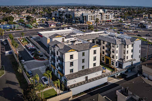 A large white building with a yellow section in the middle is surrounded by a parking lot.