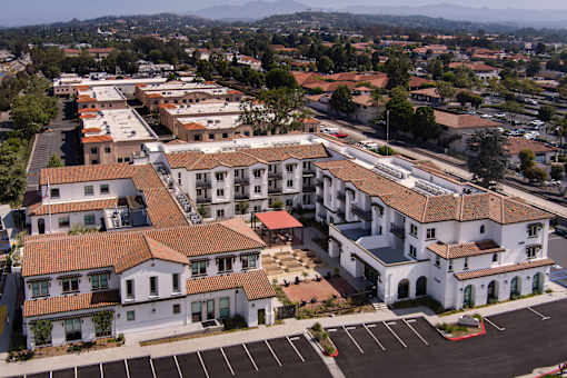 A large white building with a red roof is surrounded by other buildings and a parking lot.