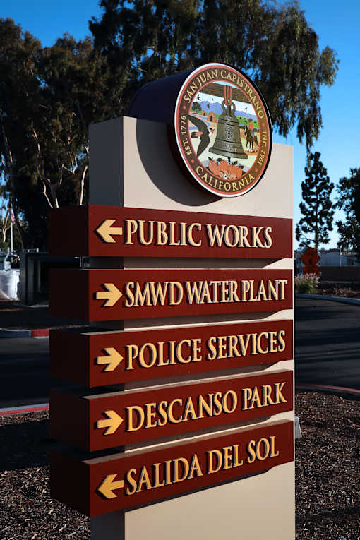 A signpost with multiple directional arrows pointing to various destinations such as Public Works, SMWD Water Plant, Police Services, Descanso Park, and Salida Del Sol.