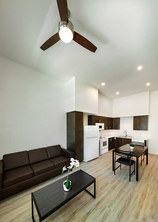 A brown couch sits in a living room with a ceiling fan and a dining table with chairs.