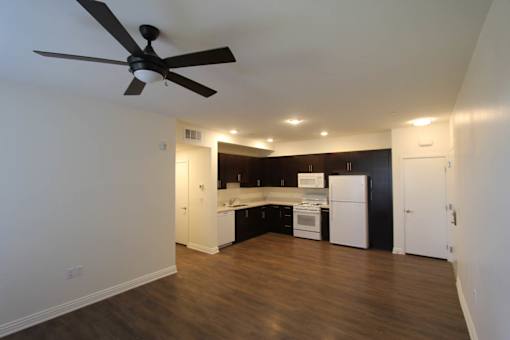 A room with a ceiling fan and a kitchen area in the background.