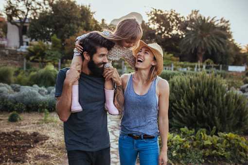 A man and woman are standing in a garden, with the man holding a child who is being lifted into the air by the woman.