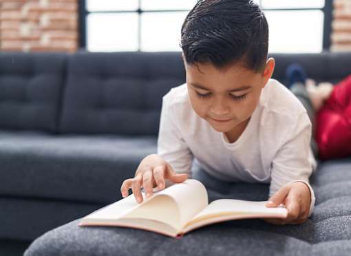 A young boy is reading a book on a grey couch.