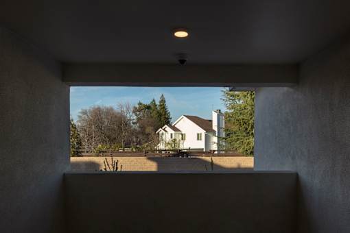 A white house is seen through a window in a room with concrete walls.