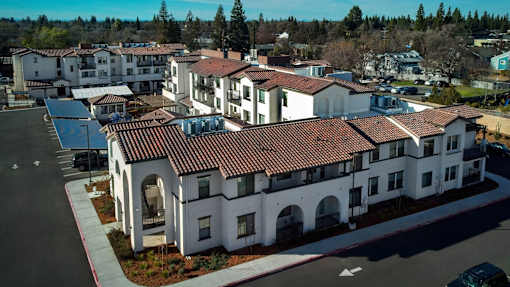 A large white building with a red tile roof is surrounded by smaller buildings and trees.