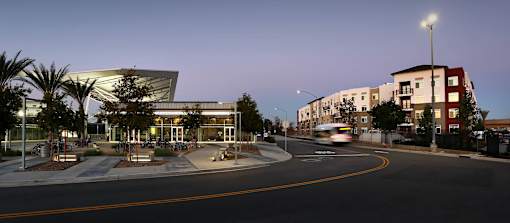 A modern building with a curved glass facade is illuminated from within, with a street in front of it.