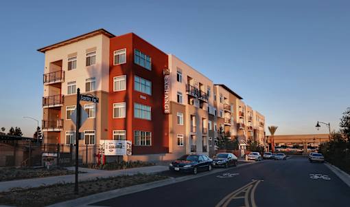 A street view of a residential area with apartment buildings and cars parked on the side of the road.