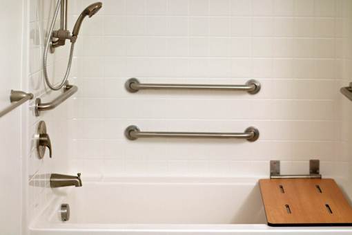 A white tiled bathroom with a shower head, hand held shower, hand held faucet, and grab bars.