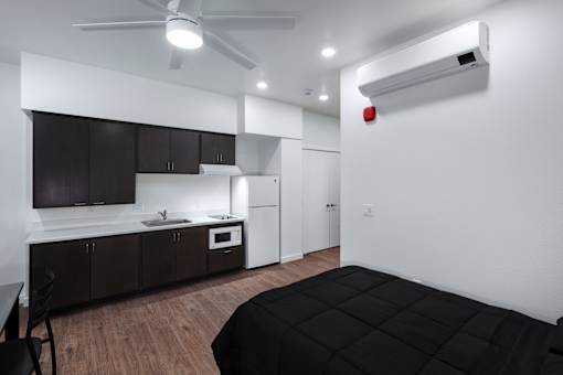 A black bed is in a white room with a kitchen area in the background.