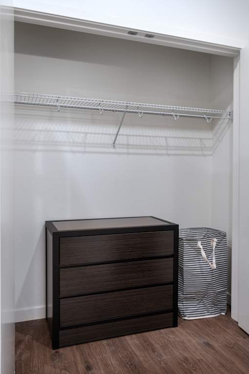 A brown chest of drawers sits next to a white bag in a room with a white wall and a white shelf.