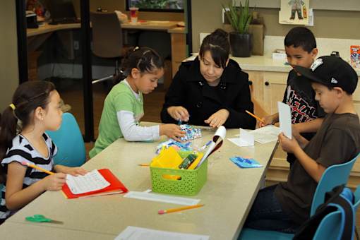 A woman is helping a young girl with her schoolwork at a table with other children.