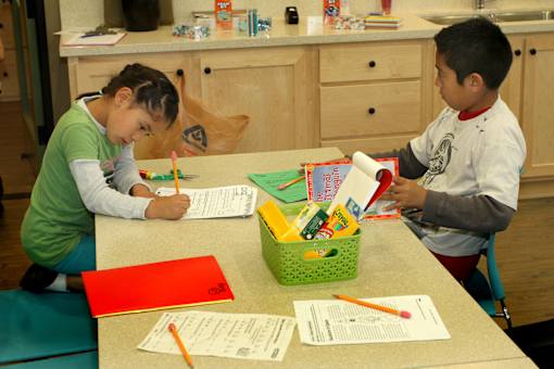 Two children are sitting at a table with school supplies and papers.