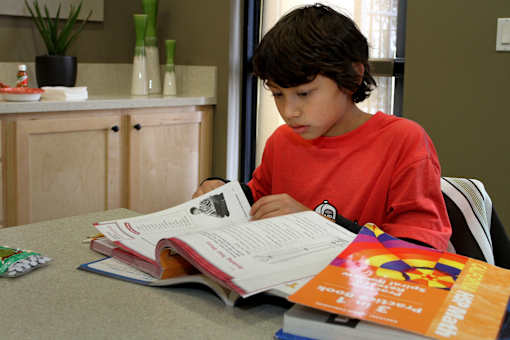 A young boy in a red shirt is reading a book at a table.