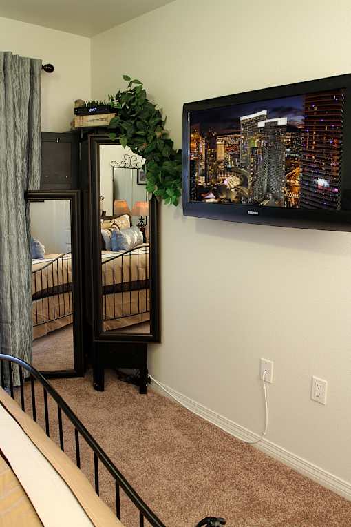 A black TV is mounted on a wall above a bed with a grey curtain.