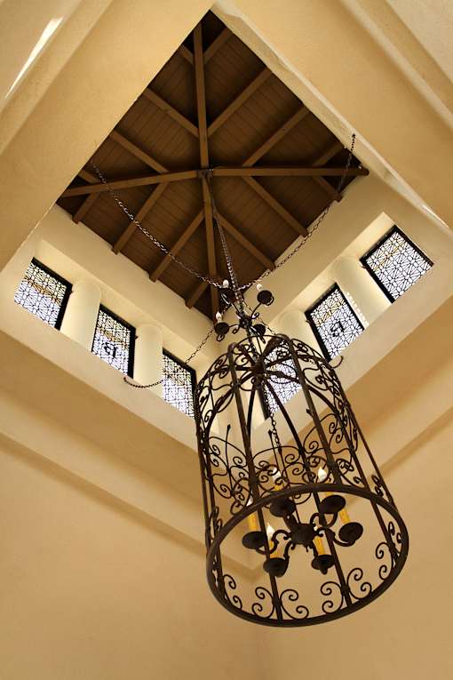 A chandelier hangs from a ceiling with a wooden beam and three windows.