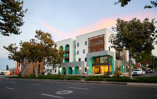 A modern building with a white facade and green accents is surrounded by trees and parked cars.