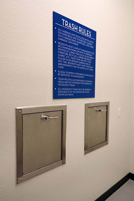 A blue sign with white text titled Trash Rules is on a wall above two silver trash receptacles.