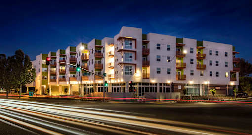 A modern apartment building is lit up at night with motion blurred street lights in the foreground.