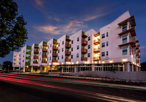 A modern apartment building with a white facade and balconies is illuminated at dusk.