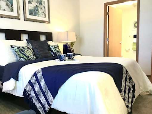 A room with a bed, nightstand and lamp with door open to bathroom.