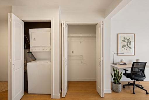 A small room with a white fridge, a black chair, a desk with a lamp and a potted plant.
