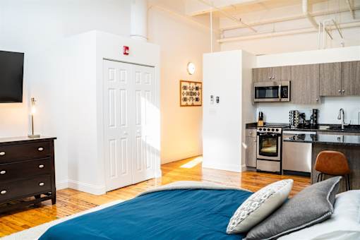 A bedroom with a bed, dresser, and a kitchen area in the background.