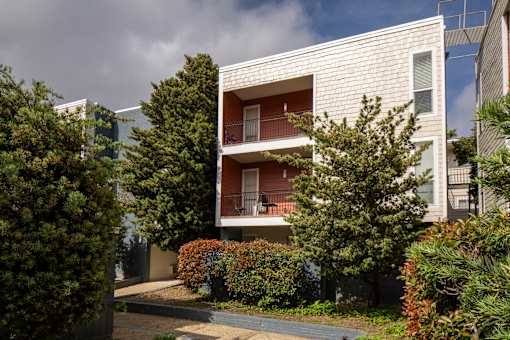 A white building with balconies is surrounded by greenery.