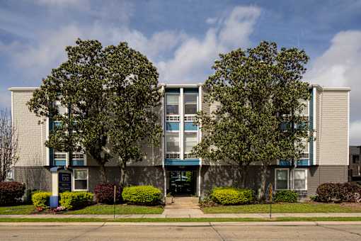 A building with a grey facade and a blue awning is surrounded by green bushes and trees.