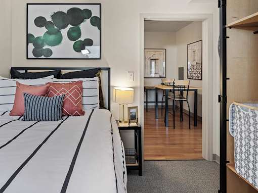 A bedroom with a bed, a nightstand with a lamp, a dining table with chairs, and a painting on the wall.