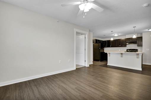 A room with a ceiling fan and a kitchen area in the background.