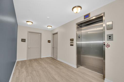 A silver elevator with a blue sign above it in a hallway with wood floors and white walls.