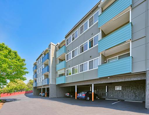 Blue, teal, and beige apartment building with carports underneath. Green trees line the left side of the paved driveway.